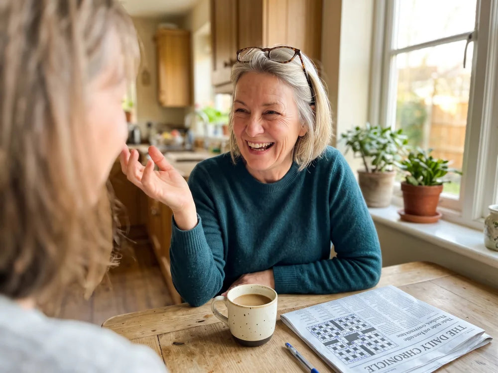 A woman in her late fifties laughing mid-story at her kitchen table, a finished crossword and coffee in front of her