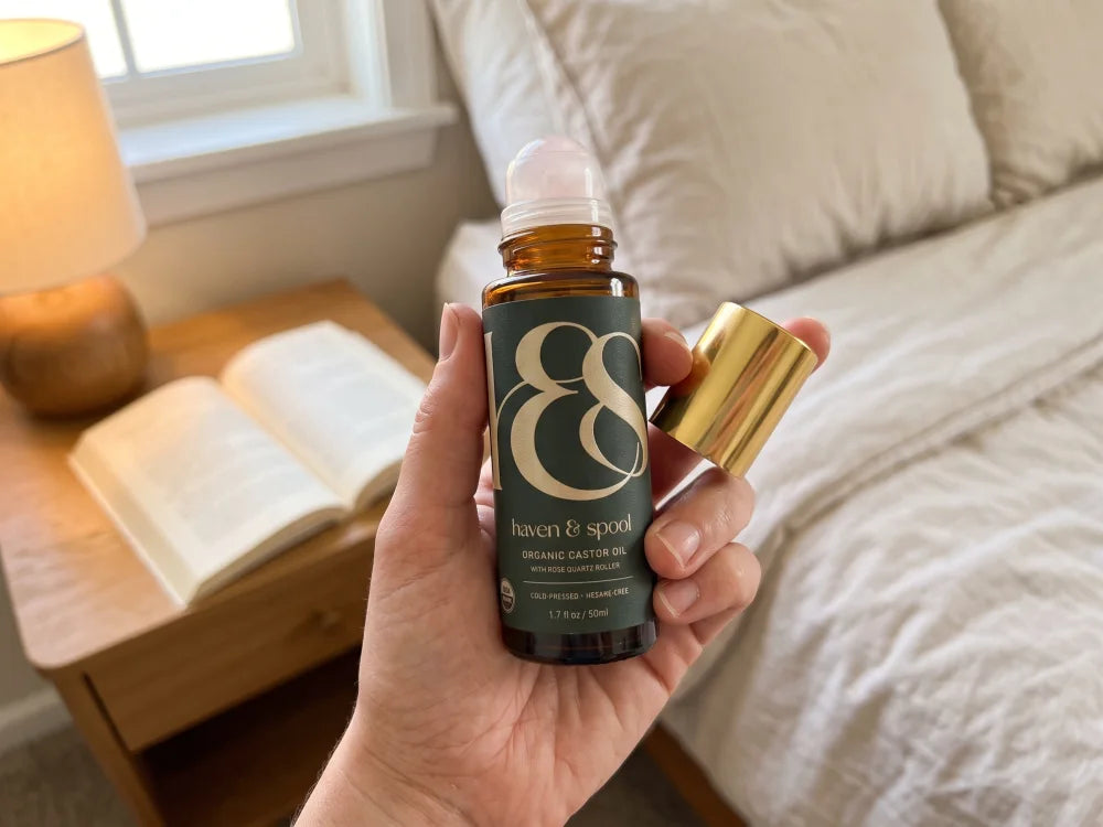 A hand holding the Haven and Spool castor oil roll-on beside a nightstand with a lamp and an open book